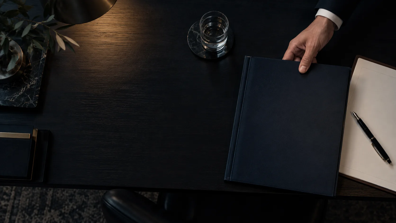 Hand opening a dark blue folder next to a notepad and pen on a desk